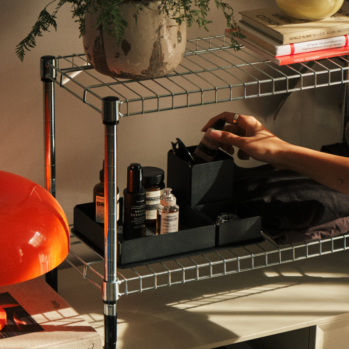 A hand reaches for a small bottle on a metal wire shelf holding various skincare products, a potted plant, and a stack of books. Warm sunlight casts shadows on the scene.