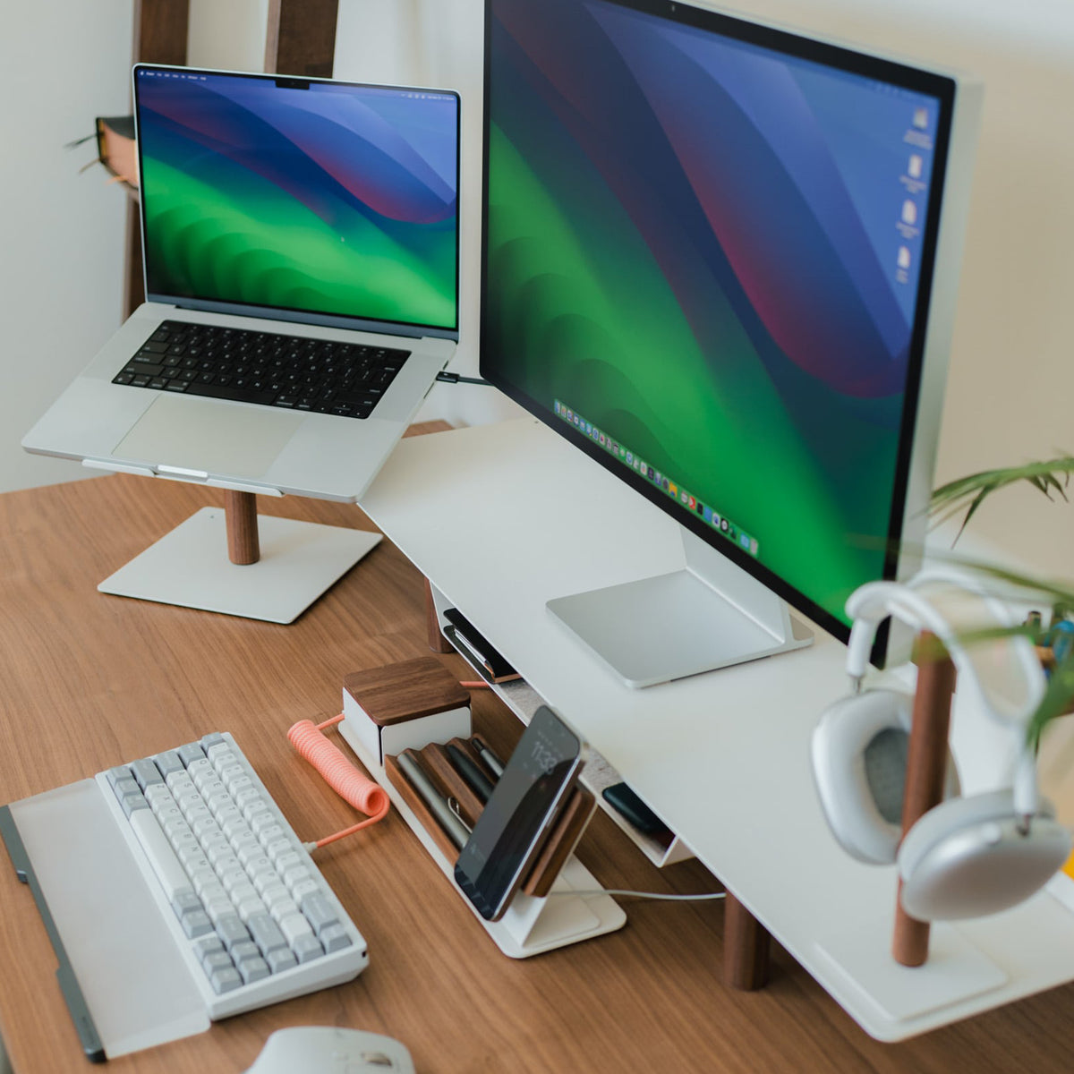 A modern workspace with a laptop and large monitor on stands, wireless keyboard and mouse, smartphone, pens, and headphones on a wooden desk with a white desk shelf organizer.