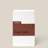 A pack of Analog Graph Cards - Small stands upright, made from uncoated cardstock with a white grid design and a brown label stating: Analog Graph Cards, 50 Cards, 3x5 in / 100 lb. The background is light beige.