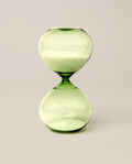 The 15 Minute Hourglass (Green), featuring green glass filled with white sand, stands upright on a simple, light background—serving as an elegant analog timer and a stylish focus tool.