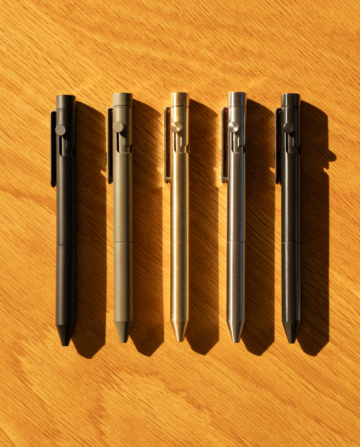Five INVENTERY Bolt Action Pens (Stainless) in black, olive green, gold, silver, and gray—each with a sleek Cerakote coating—are arranged on a light wooden surface, their metal finishes glowing in warm sunlight.