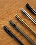 Five INVENTERY Bolt Action Pens—including a striking Zirconium edition— in black, gray, gold, silver, and black are neatly arranged diagonally on a wooden surface, their pen tips angled toward the upper right corner. Each INVENTERY Bolt Action Pen (Zirconium) exudes the refined elegance of a true luxury writing instrument.