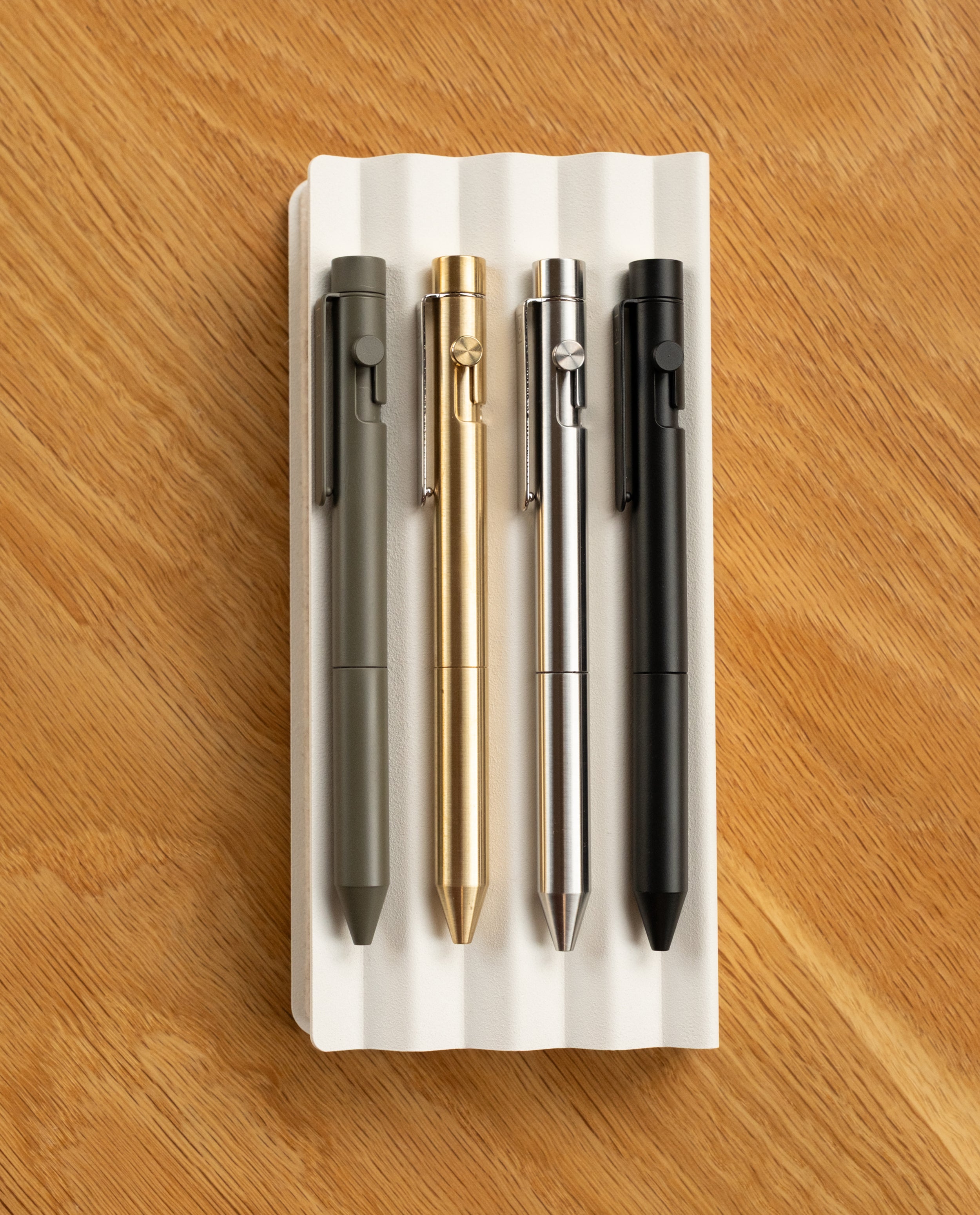Four INVENTERY Bolt Action Pens (Brass)—in green, gold, silver, and black—are arranged side by side on a white ridged tray atop a wooden surface. Each pen features a durable Cerakote ceramic coating for enhanced resilience and style.