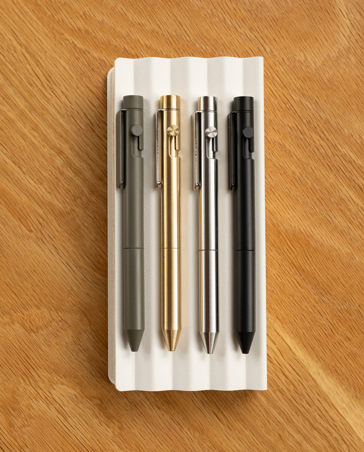 Four INVENTERY Bolt Action Pens (Stainless)—in green, gold, silver, and black—are lined up on a white grooved tray atop wood, each boasting a sleek, durable Cerakote finish.