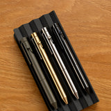 Four INVENTERY Bolt Action Pens (Stainless) in matte black, gold, silver, and black are arranged on a ridged black tray atop a wooden surface—each pen delivering style and lasting durability for daily writing tasks.