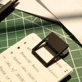 A handwritten to-do list is clipped with an HMM Clip (Set of 2 - Black) on a green cutting mat. Sunlight creates shadows, drawing attention to the list and this stylish desk organizer accessory.