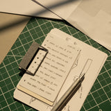 An HMM Clip (Set of 2 - Raw) secures a stack of index cards with handwritten notes and sketches, topped by a metal pen. The cards rest on a green cutting mat with grid lines, surrounded by sheets of paper.