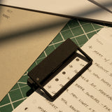 A rectangular magnifier sits on a handwritten to-do list over green and beige papers, with HMM Clips (Set of 2 - Black) and sunlight casting shadows across the organized workspace.