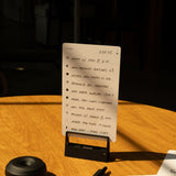 A handwritten to-do list on a small notepad sits upright on a wooden table, next to a pen, an HMM Clips (Set of 2 - Black), a round black object, and part of another notepad, all illuminated by sunlight.
