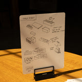 A sheet of paper, secured with HMM Clips (Set of 2 - Black), displays sketches and notes about table risers on a stand. Another sheet with handwritten notes and a black pen rests on a warmly lit wooden table.