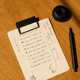 A small clipboard with a dotted notepad displays a to-do list and sketches in black ink. Nearby, HMM Clips (Set of 2 - Black) and a black pen rest on the wooden surface, sunlight casting gentle shadows across the scene.