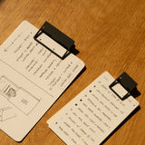 Two notepads with HMM Clips (Set of 2 - Black) lie on a wooden table—one displays a hand-drawn floor plan and notes, while the other lists tasks. A black pen, round object, and warm lighting create an organized workspace atmosphere.