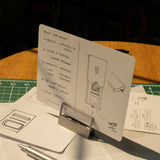 A product design sketch on a notecard rests on an HMM Clips (Set of 2 - Raw) aluminum holder, surrounded by pens, sketches, and a green cutting mat—all bathed in warm sunlight.