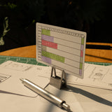 A small Gantt chart card stands on a metal holder, with scattered papers secured by HMM Clips (Set of 2 - Raw) and a silver pen on a green cutting mat, all bathed in warm sunlight.