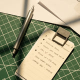 A metal pen and HMM Clips (Set of 2 - Raw) secure a handwritten to-do list on a green cutting mat, with tasks like finalizing a book collection, submitting sales tax, making calls, and sending a draft.
