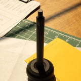 A pen stands upright in the HMM Paperweight Pen Stand (Black), a sleek desk accessory, on a desk with paper, a yellow folder, and a cutting mat. Sunlight casts shadows across the workspace. Text reads, Pen Stand sold separately.