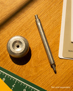 A metal HMM Paperweight Pen Stand (Raw) rests on a wooden surface near a green ruler, paint cards, and a yellow sheet. This stylish desk accessory keeps your workspace organized. Text in the corner reads, Pen sold separately.