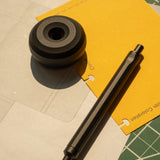 A black HMM Paperweight Pen Stand is shown on paper and a yellow folder, with sunlight casting shadows. Text in the corner reads: Pen sold separately—an ideal desk accessory for any workspace.