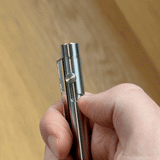 A hand holds the INVENTERY Bolt Action Pen (Stainless), thumb pressing the top button. The blurred wooden background accentuates its sleek, stainless steel design.