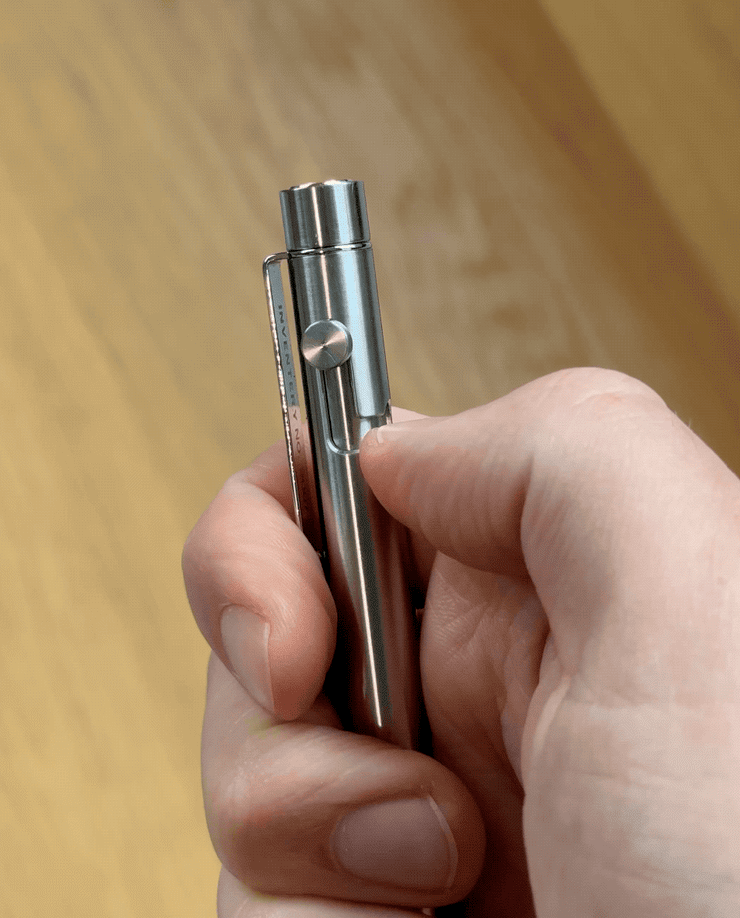 A hand holds the INVENTERY Bolt Action Pen (Stainless), thumb pressing the top button. The blurred wooden background accentuates its sleek, stainless steel design.