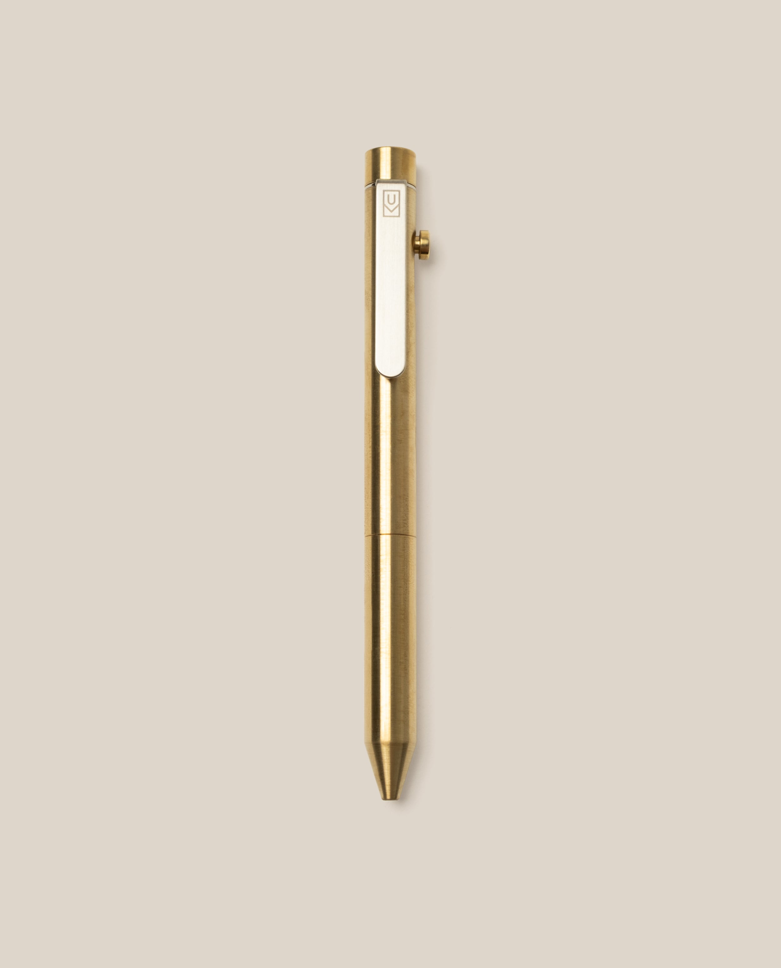 The INVENTERY Bolt Action Pen (Brass) with a clip is centered on a plain beige background. This pen showcases a sleek metallic finish and a simple, modern design, highlighting the durability of its Cerakote ceramic coating.