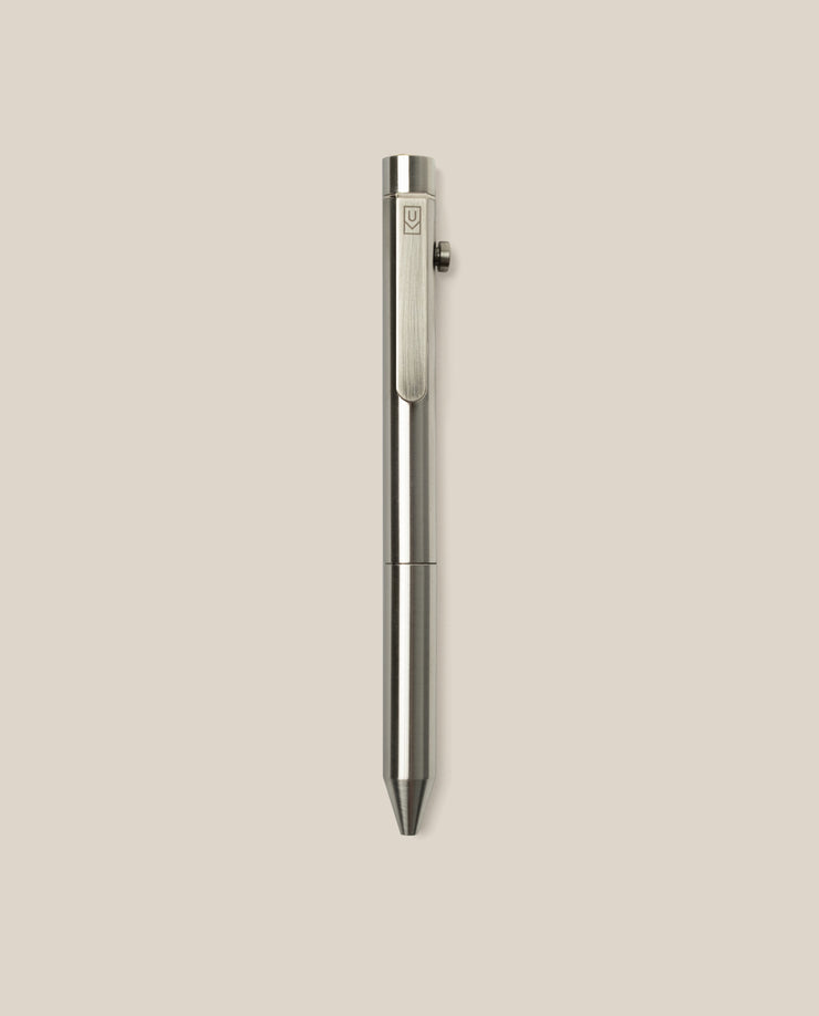The INVENTERY Bolt Action Pen (Stainless), a sleek stainless steel ballpoint with a clip, is displayed vertically against a plain light beige background.