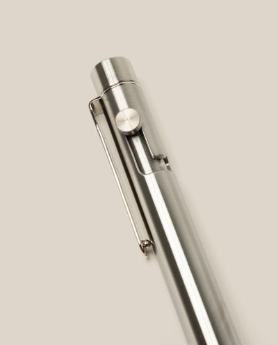 A close-up of the INVENTERY Bolt Action Pen (Stainless) with a clip, highlighting its sleek stainless steel design against a plain, light beige background.