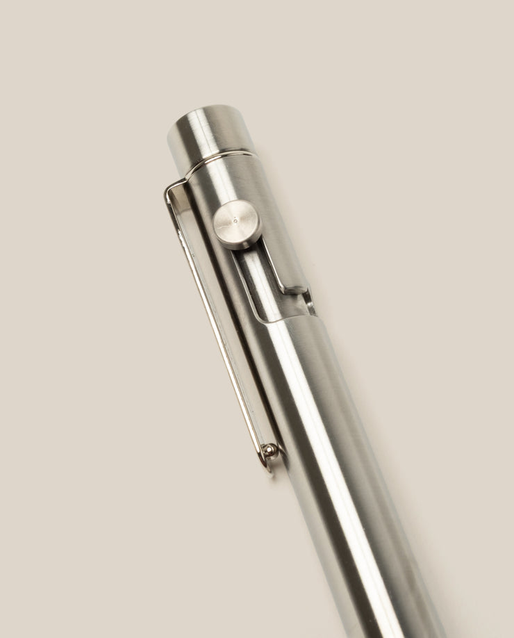 A close-up of the INVENTERY Bolt Action Pen (Stainless) with a clip, highlighting its sleek stainless steel design against a plain, light beige background.