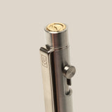 A close-up of the INVENTERY Bolt Action Pen (Stainless), featuring a gold-colored coin on top, a logo-engraved clip, and side button mechanism, set against a plain beige background.