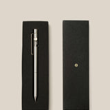 The INVENTERY Bolt Action Pen (Stainless) with a clip is displayed in an open black gift box next to its closed lid, which features a small gold emblem, all arranged on a light beige background.