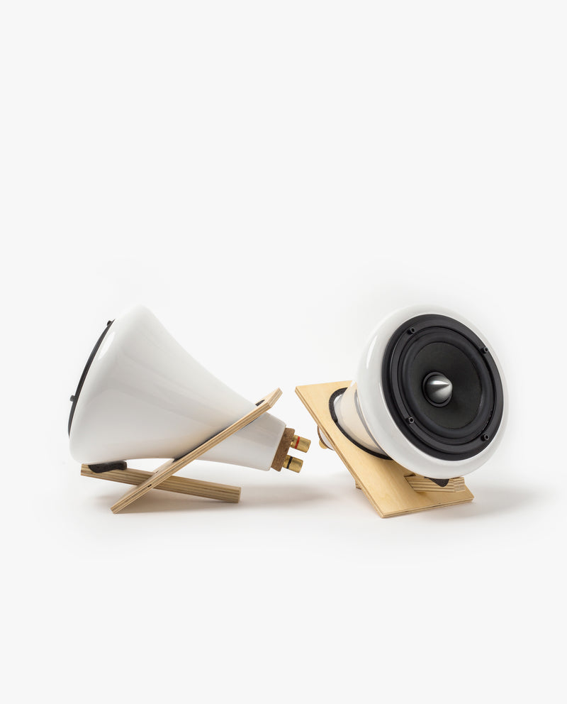 Two modern, white cone-shaped speakers with black fronts are positioned on minimalist wooden stands against a plain white background. One speaker faces forward, while the other is angled to the side.