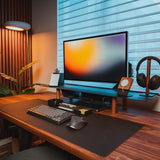 A modern desk setup features a Gather Large Monitor Stand (Black/Walnut) holding a large monitor, along with a mechanical keyboard, mouse, lamp, headphones on a stand, and plant on a wood desk by the window—an ideal space-saving workspace.