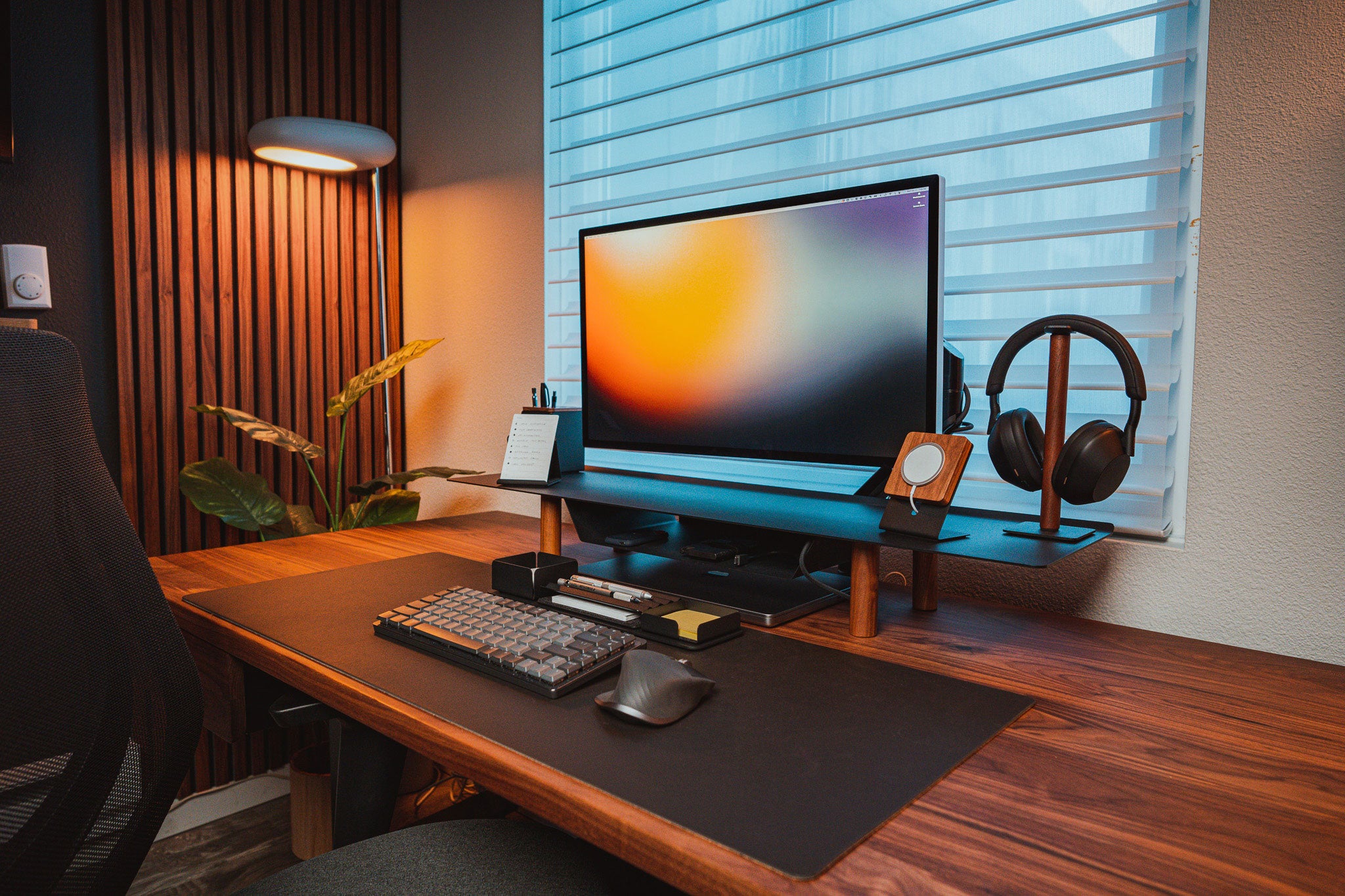 A modern desk setup features a Gather Large Monitor Stand (Black/Walnut) holding a large monitor, along with a mechanical keyboard, mouse, lamp, headphones on a stand, and plant on a wood desk by the window—an ideal space-saving workspace.