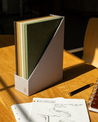 A Discbound Journal Archive with five neutral-toned notebooks sits on a wooden desk next to an open, refillable journal displaying sketches, a black pen, and a spiral-bound planner. Sunlight casts gentle shadows across the scene, creating an inviting space for creative planning and organized journal storage.