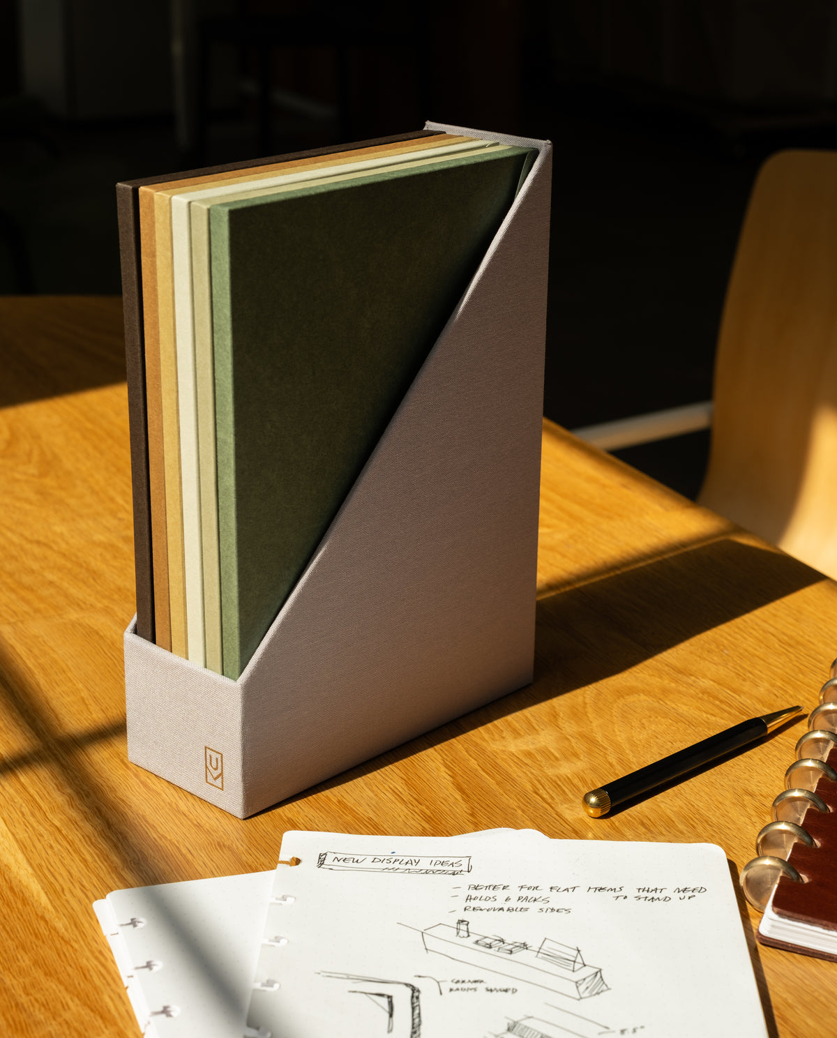 A Discbound Journal Archive with five neutral-toned notebooks sits on a wooden desk next to an open, refillable journal displaying sketches, a black pen, and a spiral-bound planner. Sunlight casts gentle shadows across the scene, creating an inviting space for creative planning and organized journal storage.