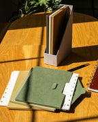 A green folder, notebooks, a pen, and a ring-bound planner are on a sunlit wooden table. Beside them lies the Discbound Heirloom Journal Bundle (Tan Leather - Standard 6″ x 8.5″) with elegant dot grid paper, while a vertical file holder with more notebooks stands in the background.