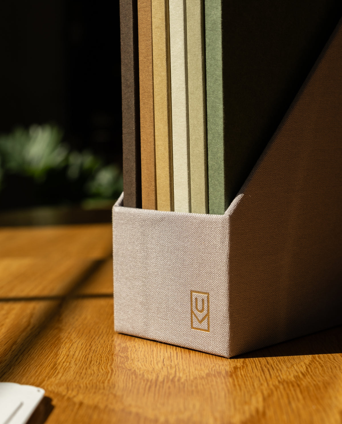 A beige file holder containing a variety of folders in brown, tan, and green rests on a sunlit wooden desk—perfect for storing or organizing your Discbound Journal Archive refills.