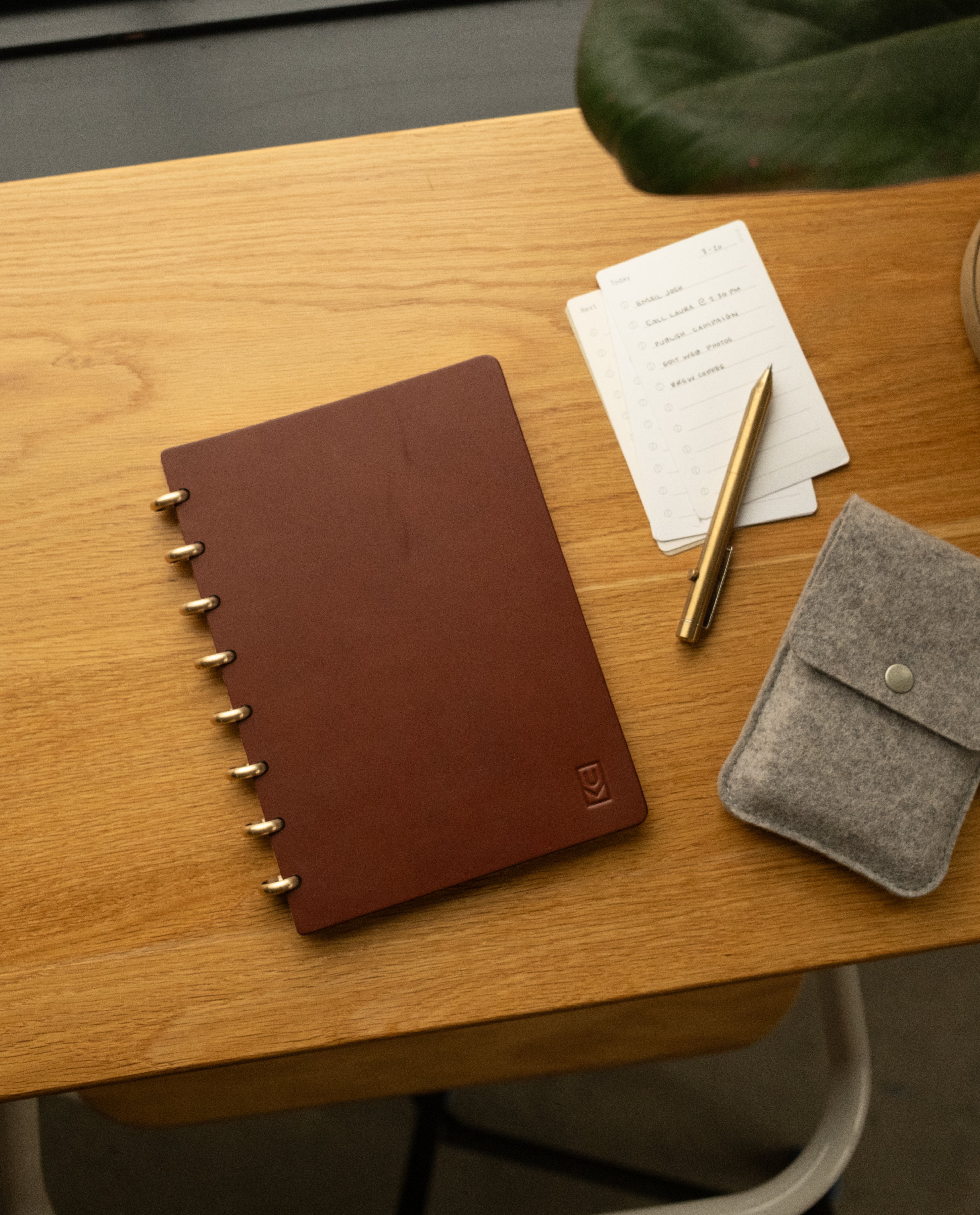 A Discbound Heirloom Journal in brown leather (6" x 8.5"), a gold pen, dot grid note cards with to-do list, and a gray felt pouch are arranged on a wooden desk beside a green plant.
