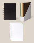 A Discbound Heirloom Journal Bundle in black leather (Standard 6″ x 8.5″) is displayed with four loose dot grid paper pages and a gray holder containing five full-grain leather journals in assorted earth tones, all set against a beige background.