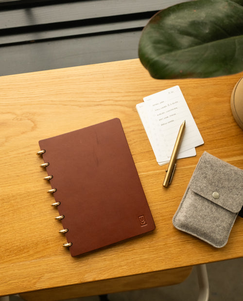 A Discbound Heirloom Journal in brown leather (6″ x 8.5″) with three refills, a gold pen, note cards, and a gray felt pouch rest on a wooden desk near a potted plant.