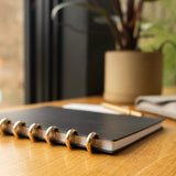 A Discbound Heirloom Journal Bundle (Black Leather - Standard 6" x 8.5" + 3 Refills) sits closed on a wooden desk beside a potted plant, with a pen and gray cloth partially visible nearby in natural window light.