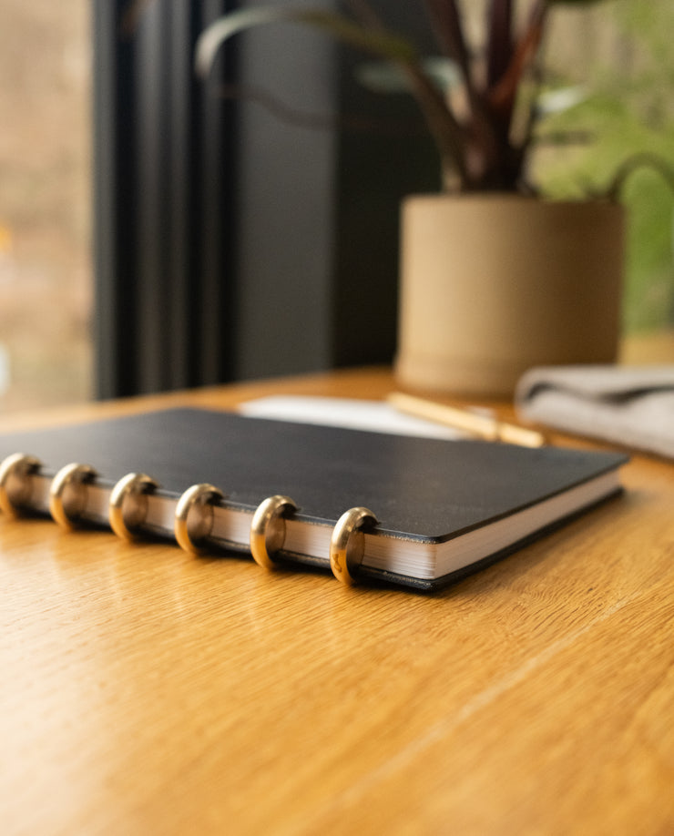 A Discbound Heirloom Journal Bundle (Black Leather - Standard 6" x 8.5" + 3 Refills) sits closed on a wooden desk beside a potted plant, with a pen and gray cloth partially visible nearby in natural window light.