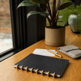 A Discbound Heirloom Journal (Black Leather - Standard 6" x 8.5"), a pen, and a small pouch sit on a wooden desk near a window, softly illuminated by sunlight with a potted plant in the background.