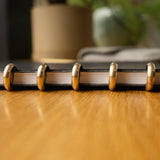A close-up side view of the Discbound Heirloom Journal (Black Leather - Standard 6" x 8.5") with gold disc rings resting on a wooden table, blurred objects in the background.