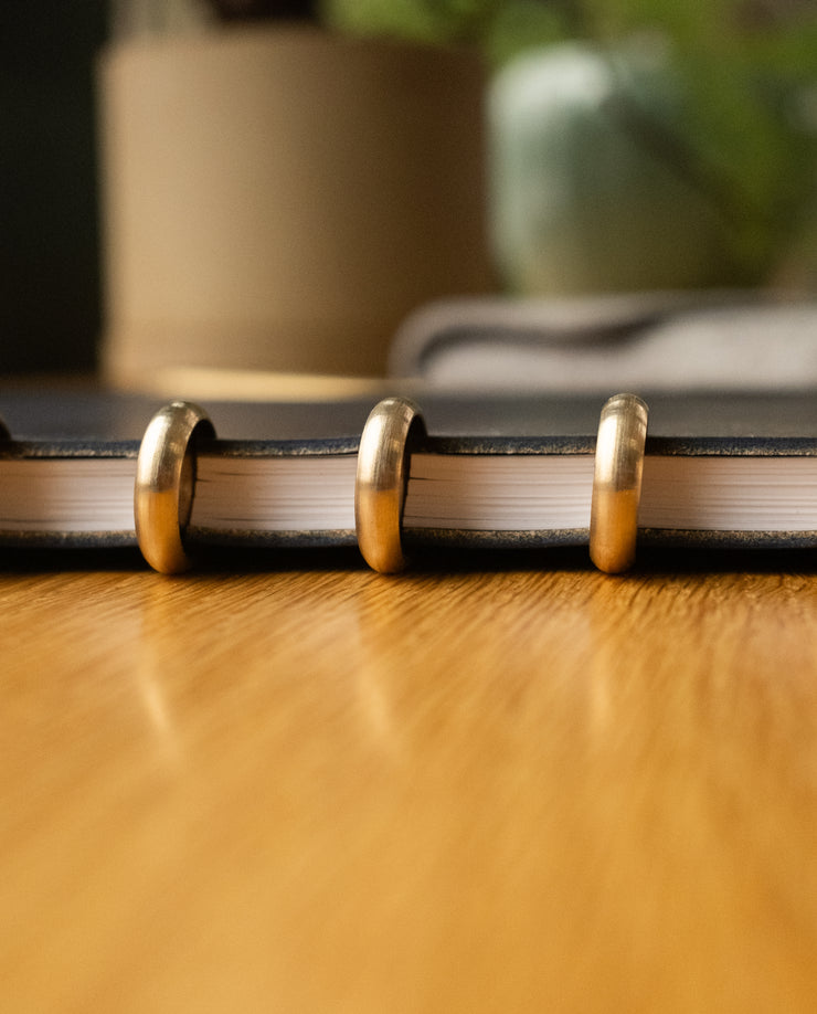 A close-up side view of the Discbound Heirloom Journal (Black Leather - Standard 6" x 8.5") with gold disc rings resting on a wooden table, blurred objects in the background.