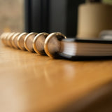 A Discbound Heirloom Journal (Black Leather - Standard 6" x 8.5") lies on a wooden surface, showcasing its durable metal rings in sharp focus against a softly blurred background.