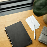 A Discbound Heirloom Journal Bundle (Black Leather - Standard 6" x 8.5" + 3 Refills) sits on a wooden desk near a window, accompanied by a silver pen, lined note cards, part of a potted plant, and a gray felt pouch.