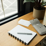 A silver pen rests on a black leather Discbound Heirloom Journal Bundle (Standard 6" x 8.5" + 3 Refills) on a wooden desk near a window, with a potted plant, felt pen case, and note cards in sunlight.