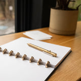 A Discbound Heirloom Journal (Black Leather - Standard 6" x 8.5") and a gold pen rest on a wooden desk by a window, with a potted plant and small stack of cards in the background.