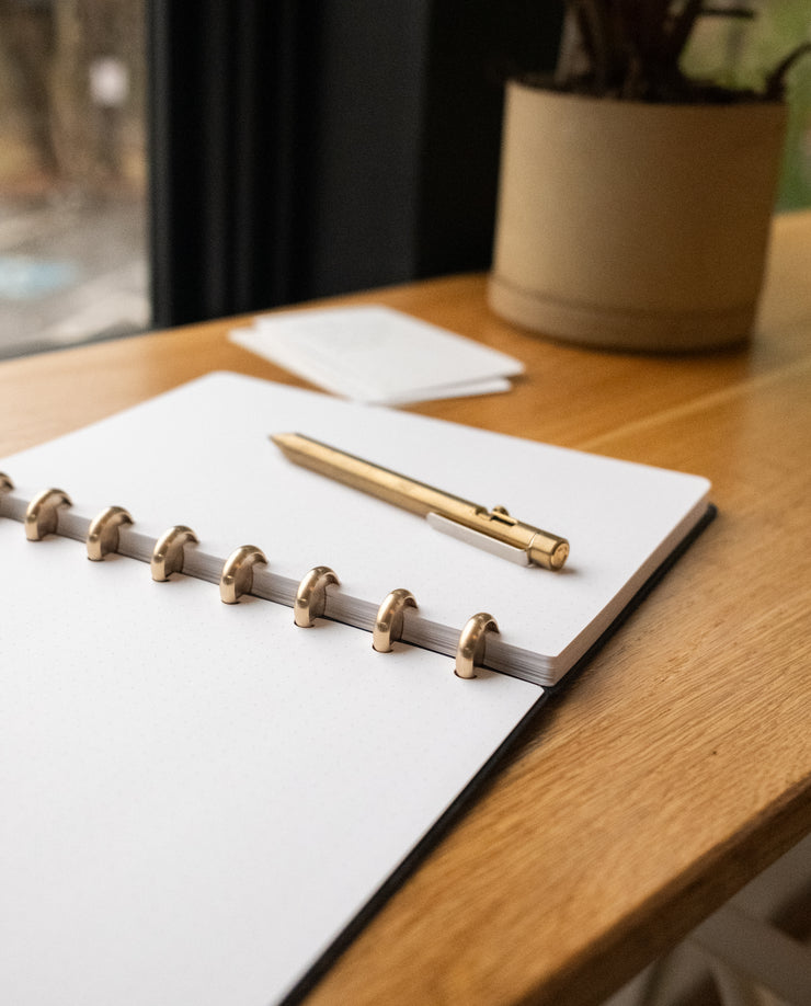A Discbound Heirloom Journal (Black Leather - Standard 6" x 8.5") and a gold pen rest on a wooden desk by a window, with a potted plant and small stack of cards in the background.